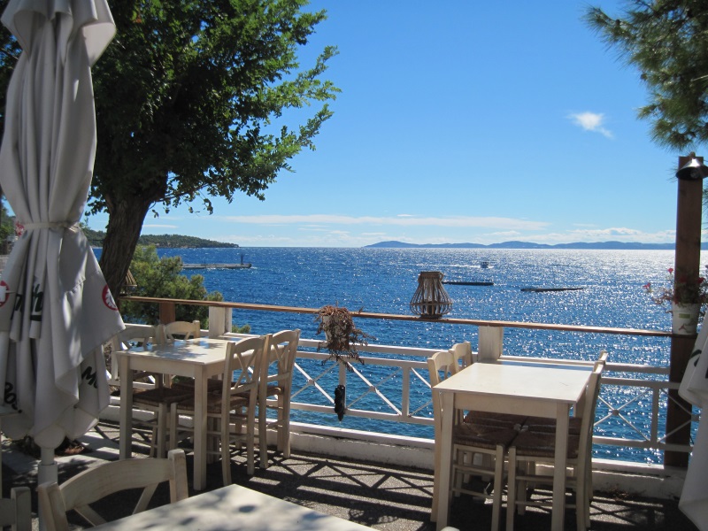 Terrace of a Greek tavern by the sea around noon, overlooking sparkling waves, fishing boats, and a distant peninsula.