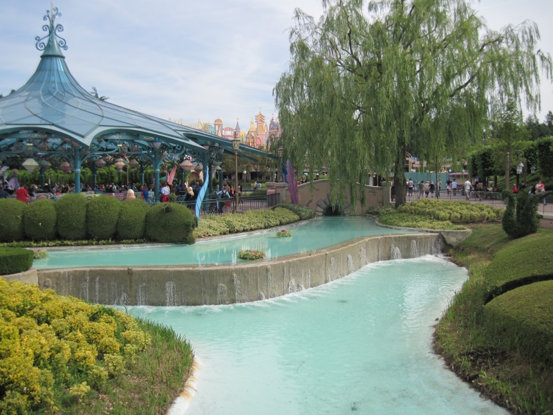 A section of Fantasyland in Disneyland Park, Paris, features blue ponds edged with greenery and an entertainment pavilion.
