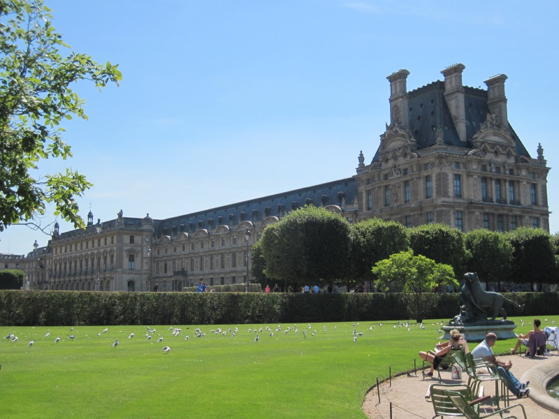 View of Louvre Museum's Denon Wing from Tuileries Garden, with a tree line and a lush green fence standing between them.