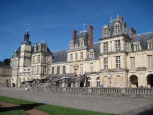 Front façade of Chateau de Fontainebleau, a French royal palace, with a horseshoe-shaped staircase as seen on a sunny day.