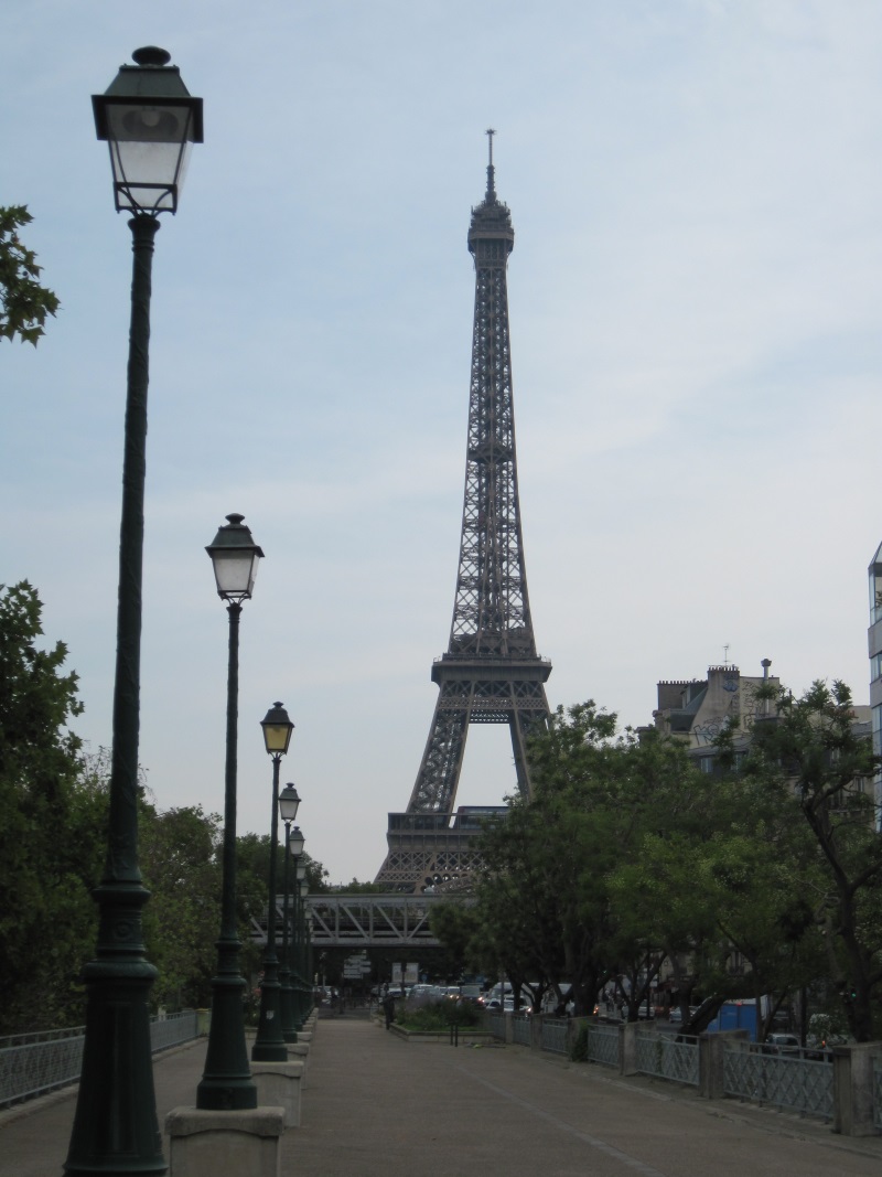 Eiffel Tower dominates the Paris skyline while a walking path lined with trees and street lanterns leads to the monument.