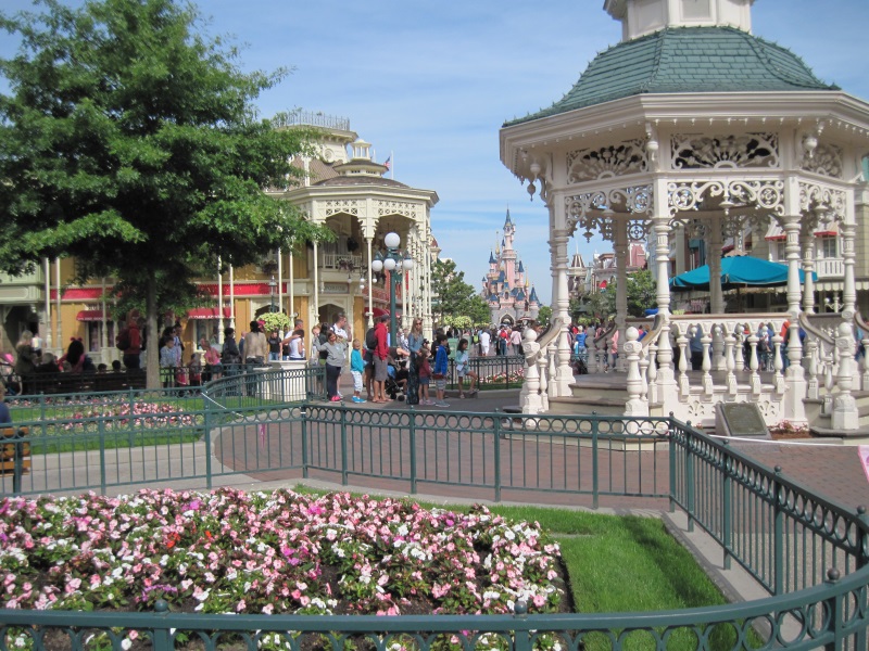 View of a pavilion, flower beds, commercial buildings, and distant Sleeping Beauty's Castle in Disneyland Park, Paris.