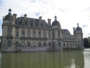 View of the Renaissance Château de Chantilly with semi-oval cupolas flanked by a canal with a cloudy sky in the background.