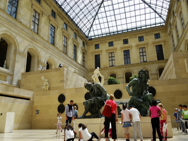 Louvre Museum visitors examine dramatic sculptures, one of which is Milo of Croton by Pierre Puget, in a well-lit courtyard.