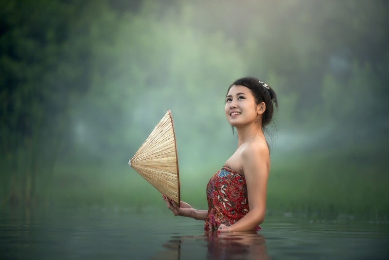 A smiling young Cambodian lady, holding a traditional conical hat, poses for a photo in a shallow pond in a lush setting.