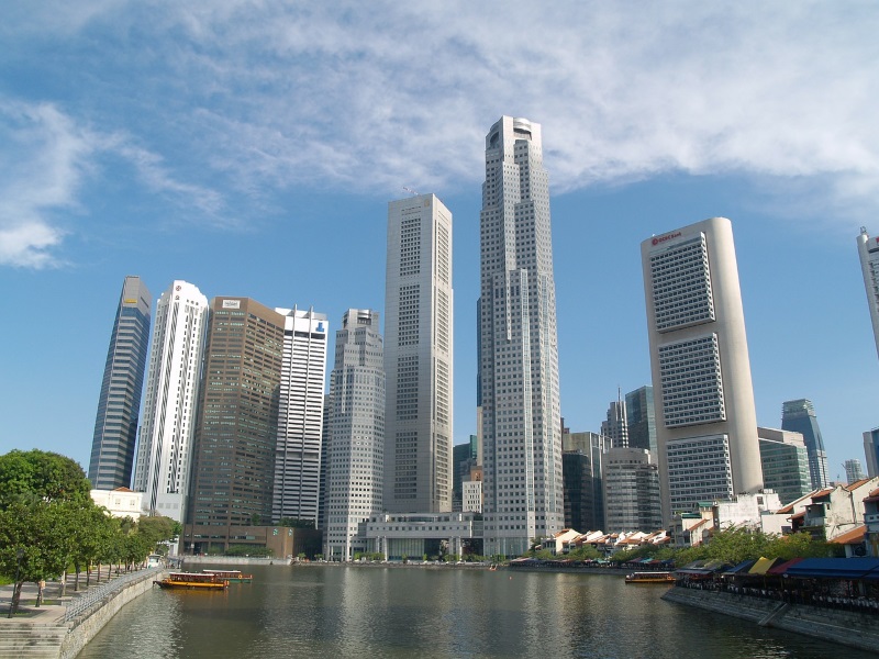 Modern skyscrapers define the Singapore River's skyline while trees line its western shore and boats navigate the river.